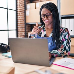 African woman with braids working at small business ecommerce with laptop smiling happy and positive, thumb up doing excellent and approval sign